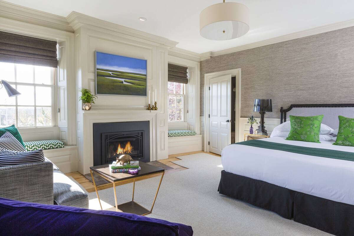 Une chambre avec une cheminée, un grand lit avec des coussins verts, un canapé et un tableau au mur.