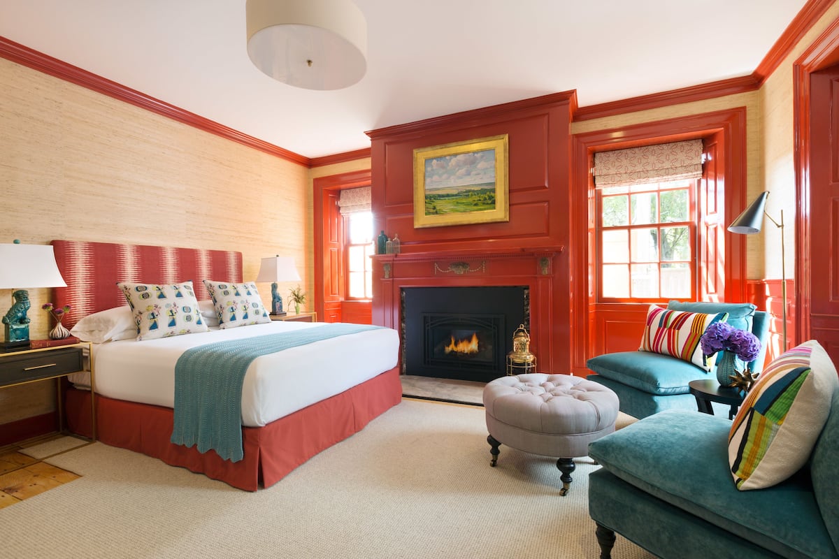 Une chambre à coucher avec un grand lit, une cheminée, une fenêtre avec des rideaux et un fauteuil turquoise.