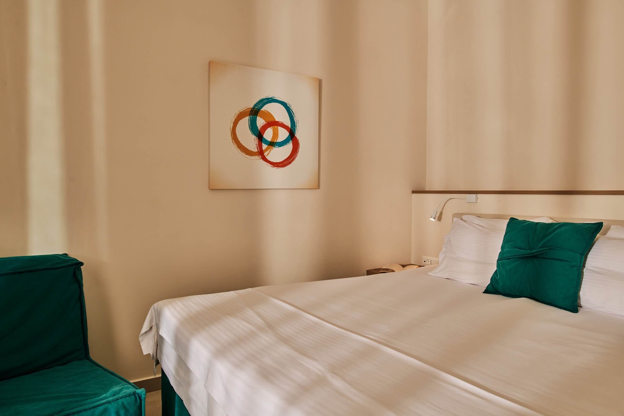 A neatly made bed with a green pillow, a wall-mounted abstract painting, and a green chair.