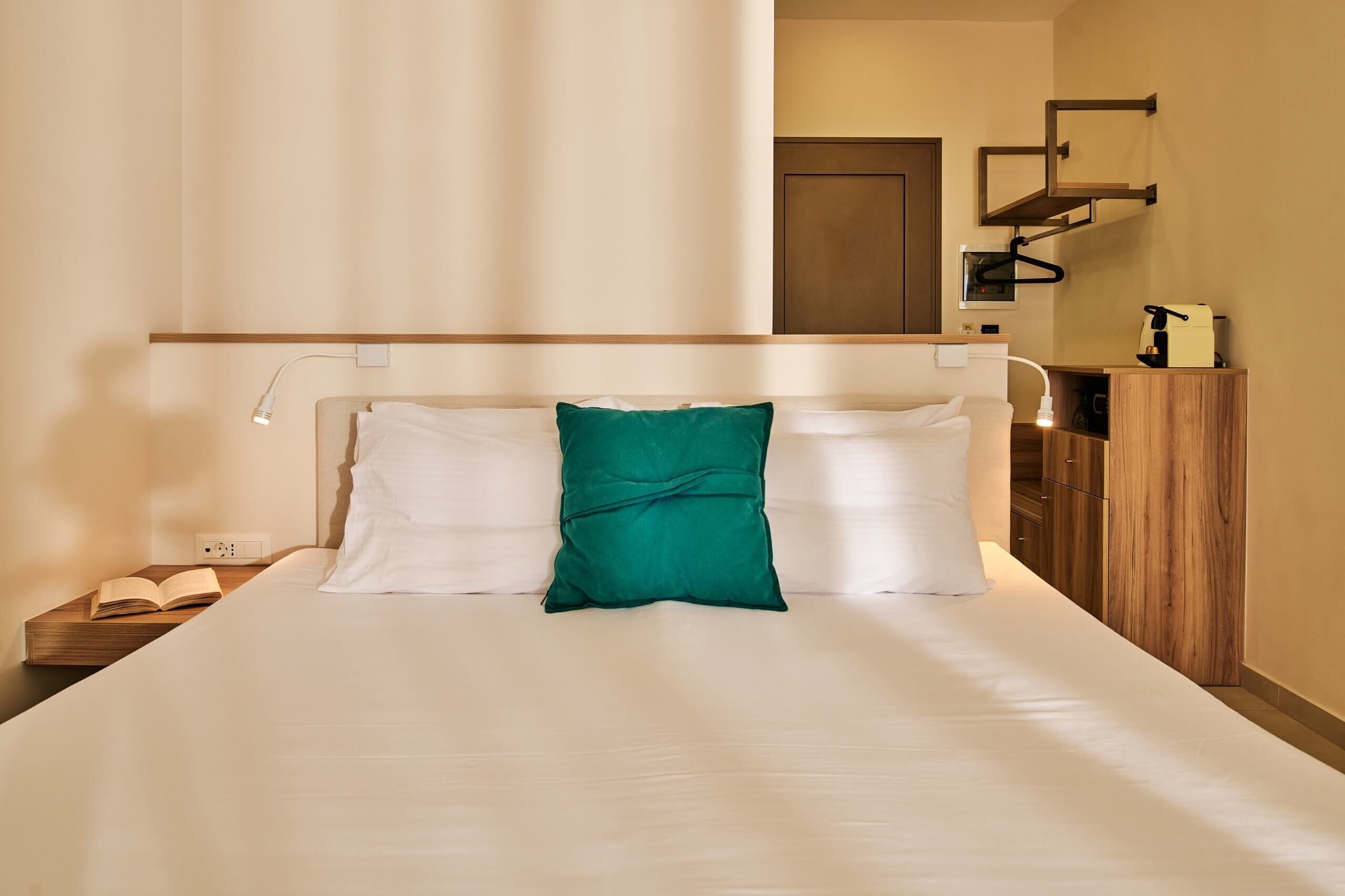 A neatly made bed with white and green pillows, a bedside table with a book, and a nightstand with a lamp.