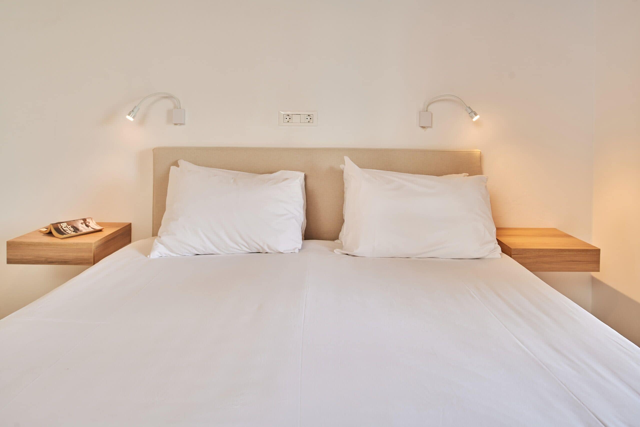 A neatly made bed with white linens, two bedside tables with lamps, and a wall socket above the bed.