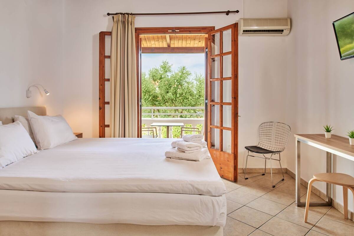 Une chambre à coucher comprenant un lit, un bureau, une chaise et une porte ouverte donnant sur un balcon avec vue sur les arbres.