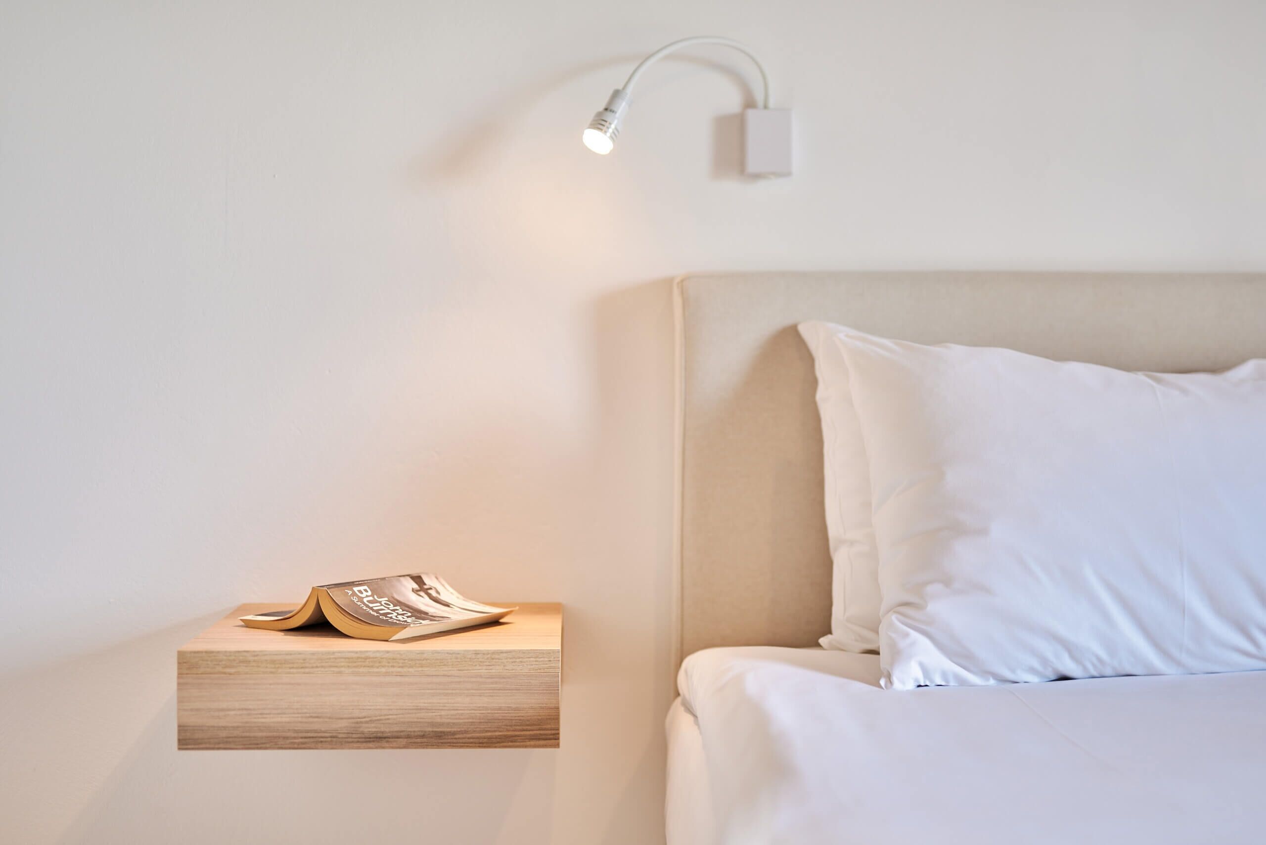 Una cama bien hecha con ropa blanca, una mesita de noche con un libro y una lámpara de pared.