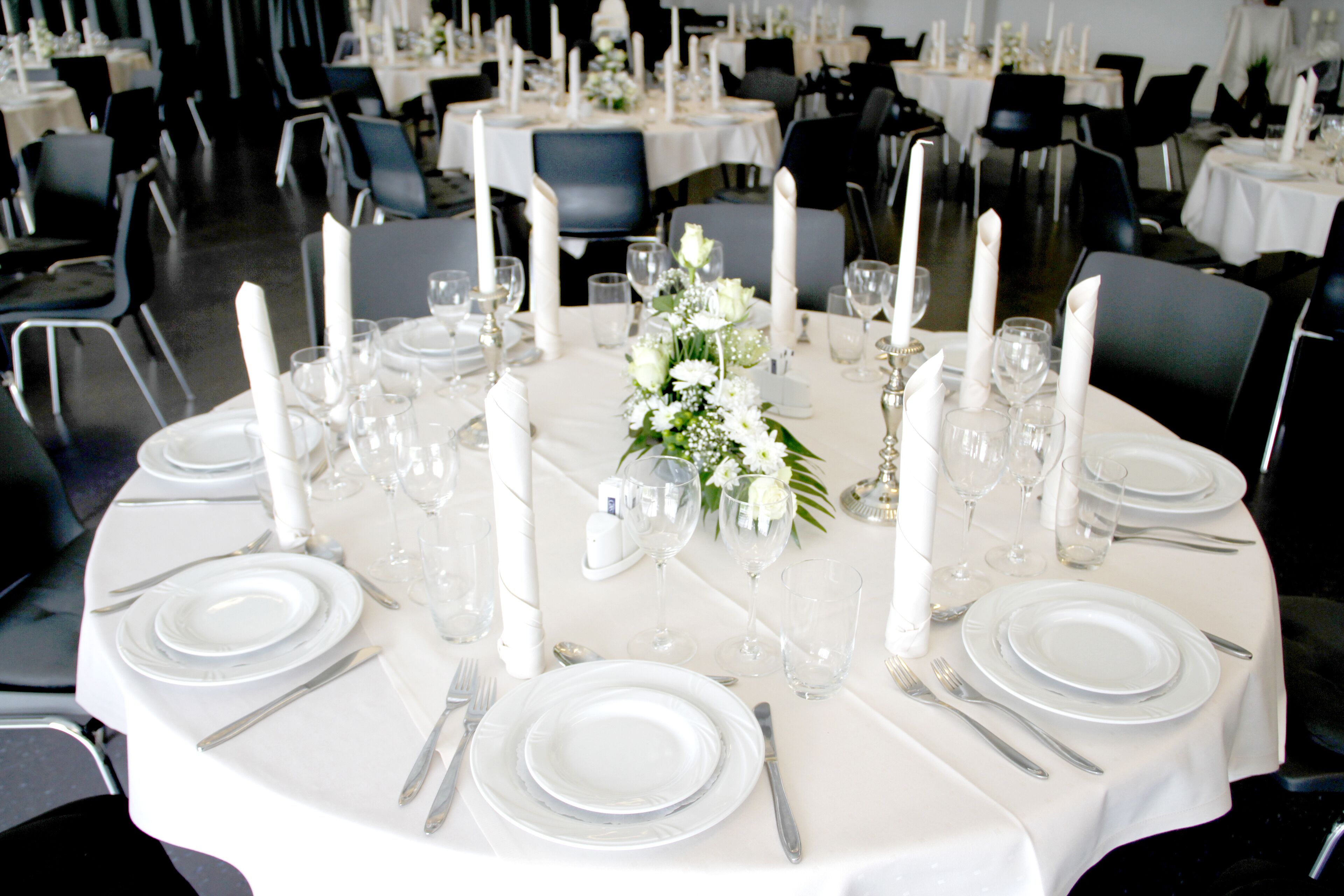 Una mesa redonda dispuesta para un evento formal, con manteles blancos, platos, cubiertos y velas.