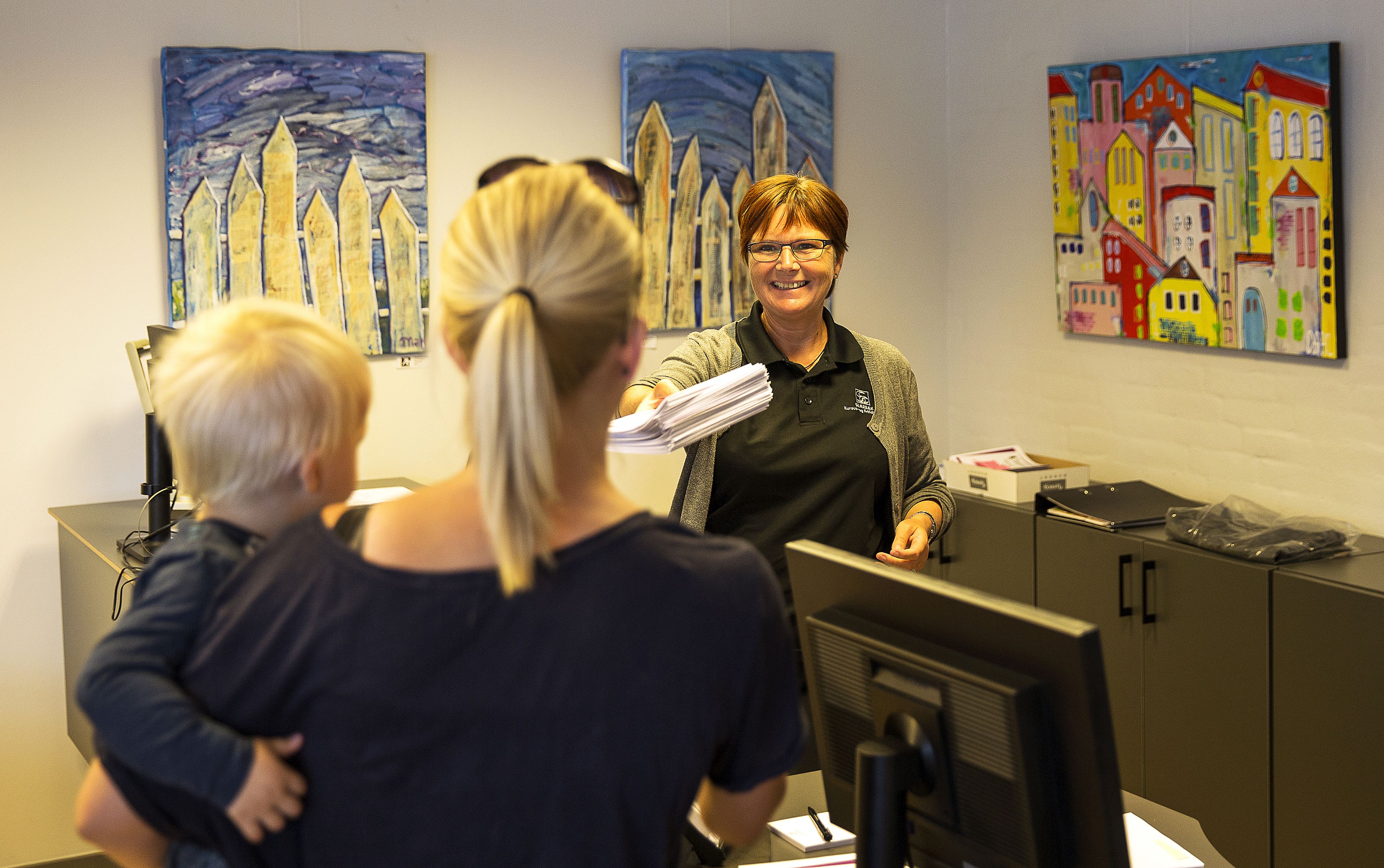 A reception desk with a computer monitor, a telephone, and a box of tissues. There are two paintings on the wall behind the desk.