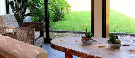 A sunlit patio with wicker furniture, a round table with potted plants, and a view of a green lawn.