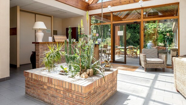 Un vaste espace intérieur doté d’un plafond en verre, d’un jardin en briques où sont plantées différentes espèces, de meubles en osier et d’une vue sur l’extérieur.