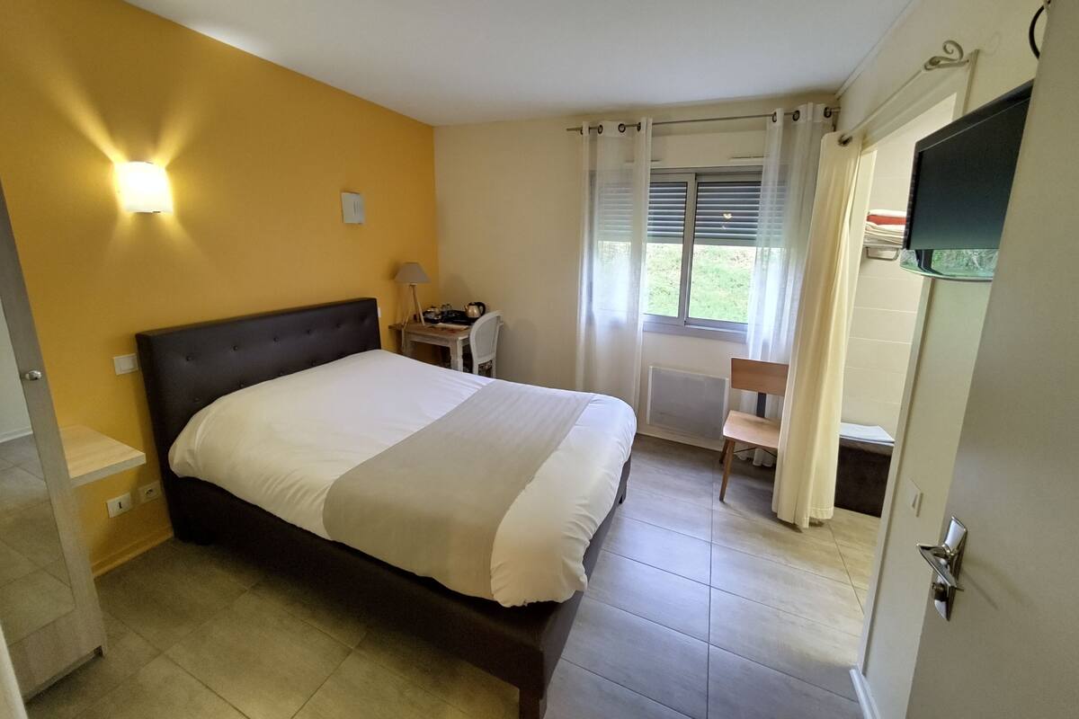 A bedroom with a bed, a desk, a chair, a window with curtains, and a television.