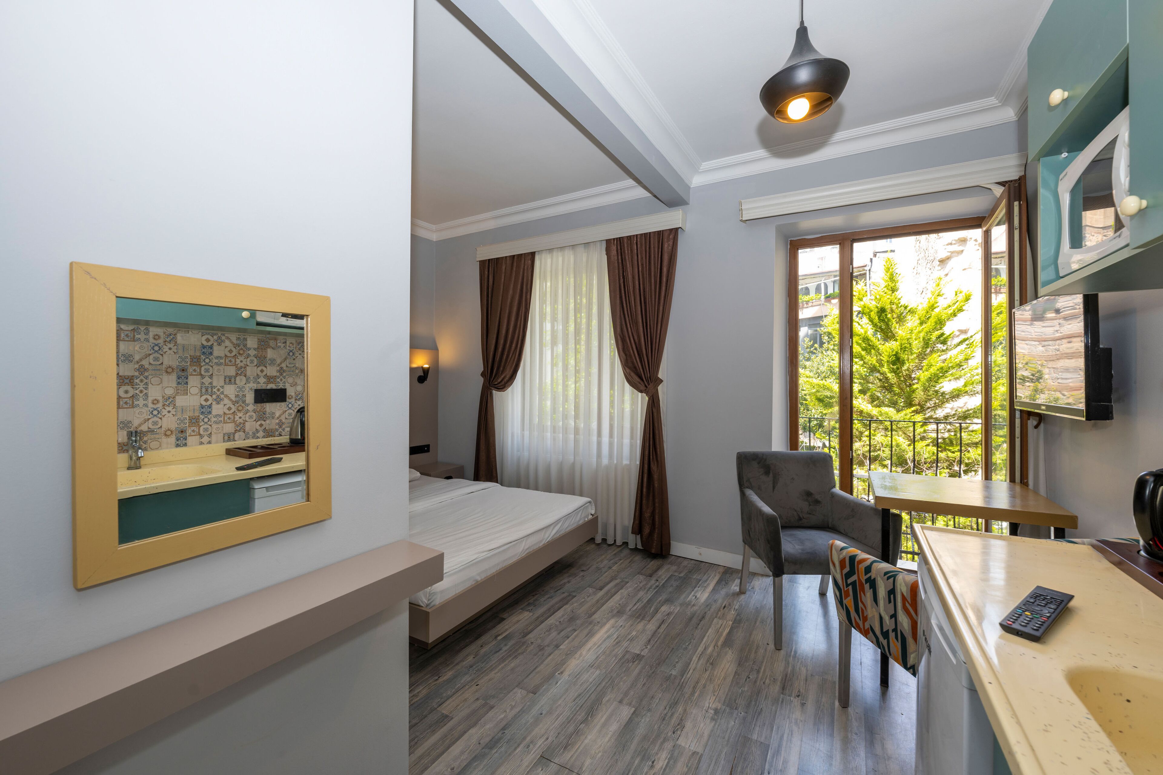 A hotel room with a bed, a desk, a chair, a window with curtains, and a view of greenery outside.