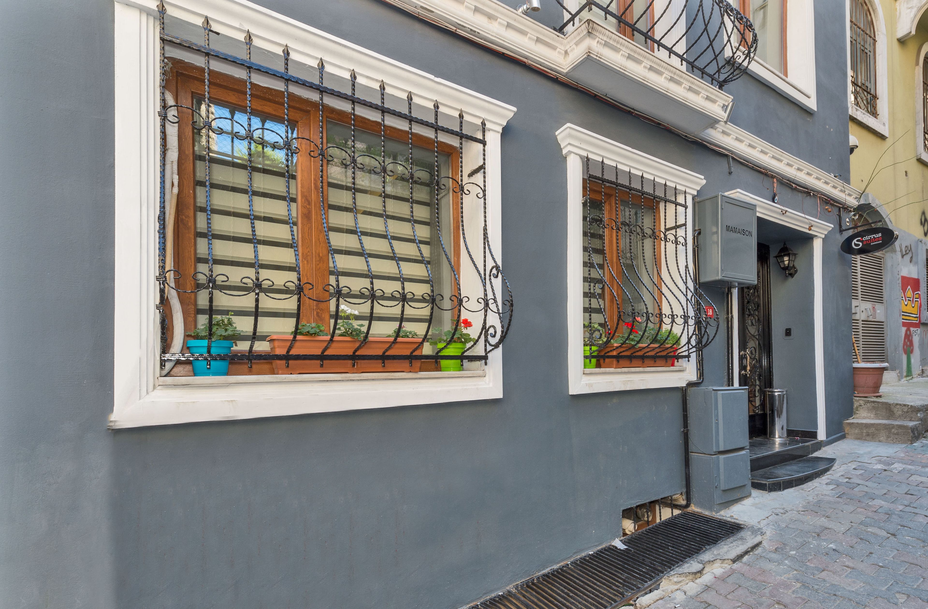 A building facade with two windows featuring wrought iron grilles and wooden shutters, potted plants on the windowsills, and a small entrance with steps.