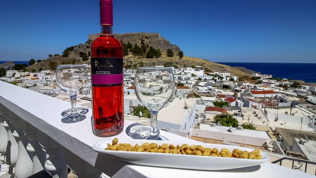 A balcony with a bottle of rosé wine, two wine glasses, and a plate of nuts against a backdrop of a coastal town and a mountain. - Domna Studios (Rhodes)