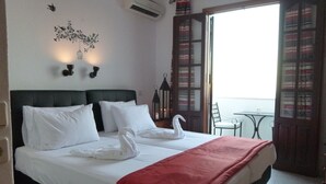 A hotel room with a bed, white pillows, a red blanket, a wall-mounted light fixture, and a view of the outside balcony. - Domna Studios (Rhodes)