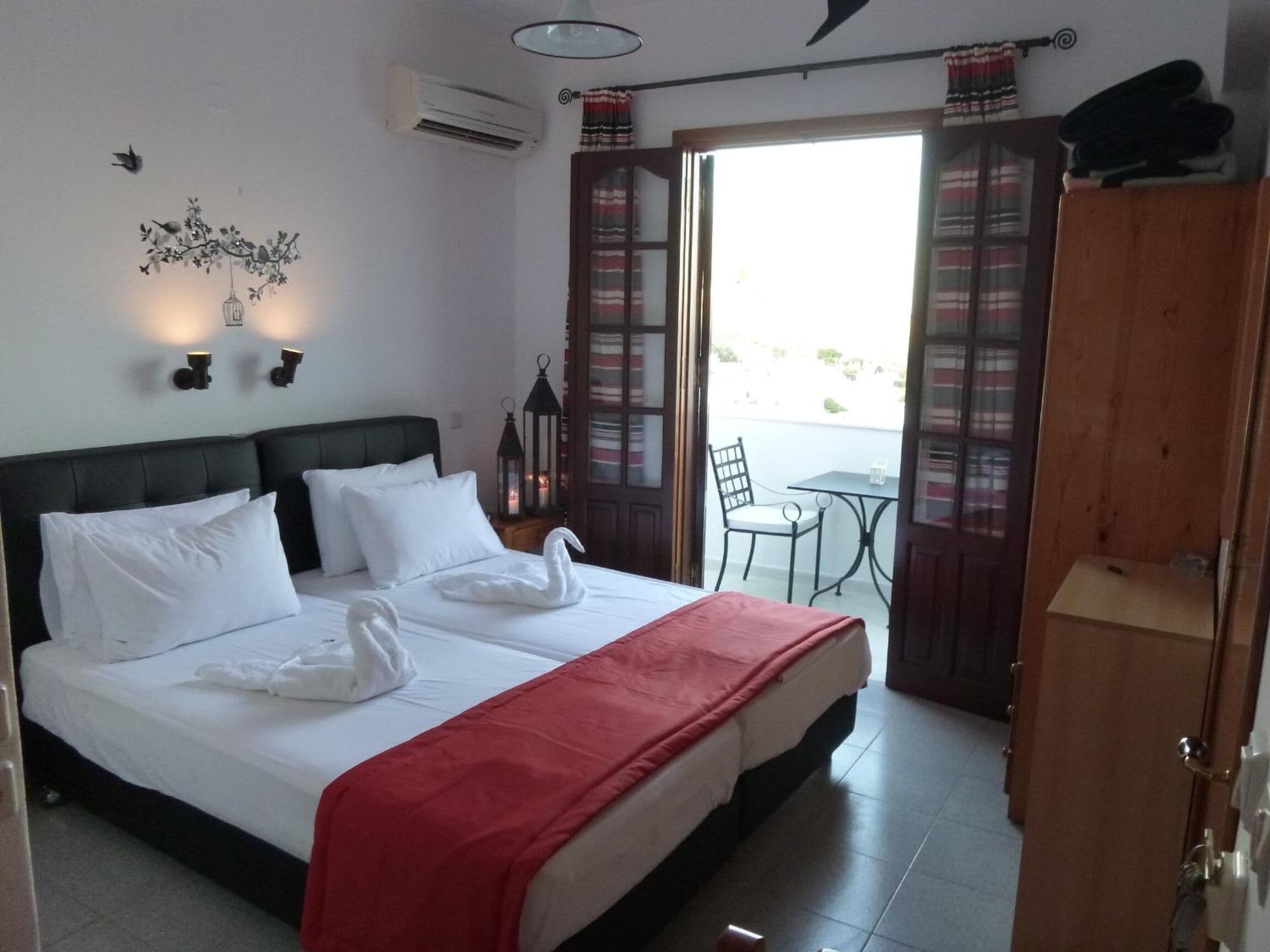 Habitación de hotel con cama, dos luces fijadas a la pared, unidad de aire acondicionado, balcón con mesa y silla, y armario de madera.