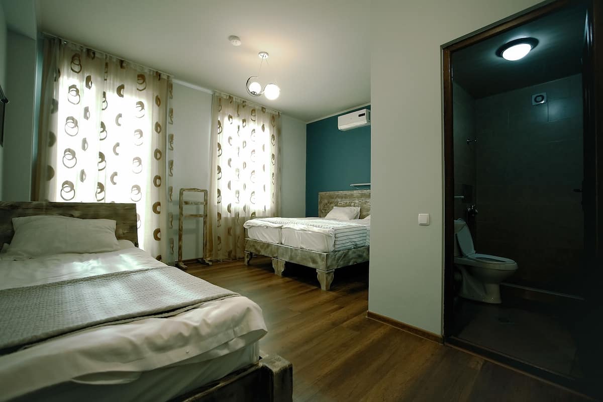 A bedroom with two beds, a wooden floor, a bathroom with a toilet and shower, and a ceiling light.