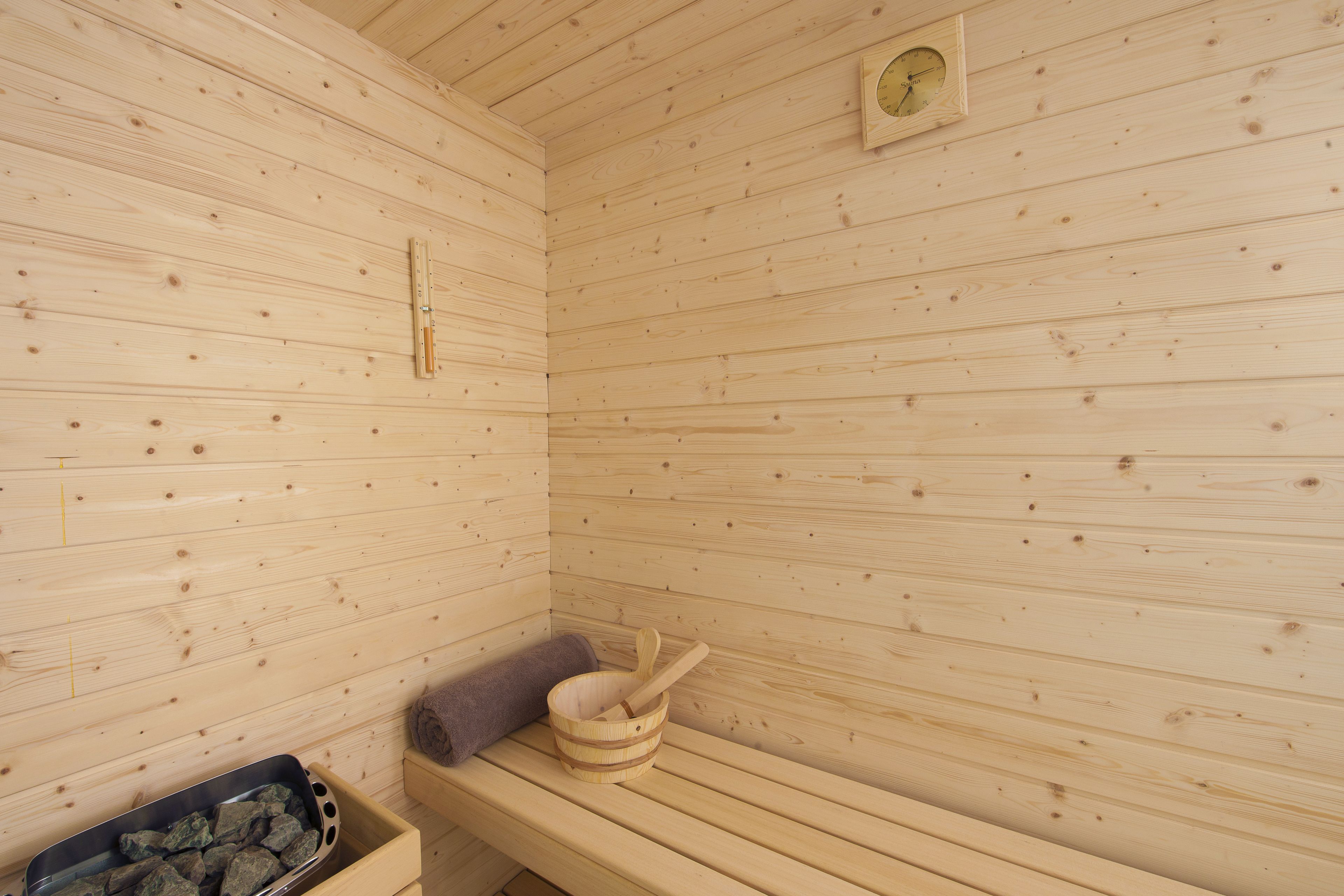 Una sauna de madera con un banco, un cubo de madera y una cuchara para servir, además de un termómetro en la pared.
