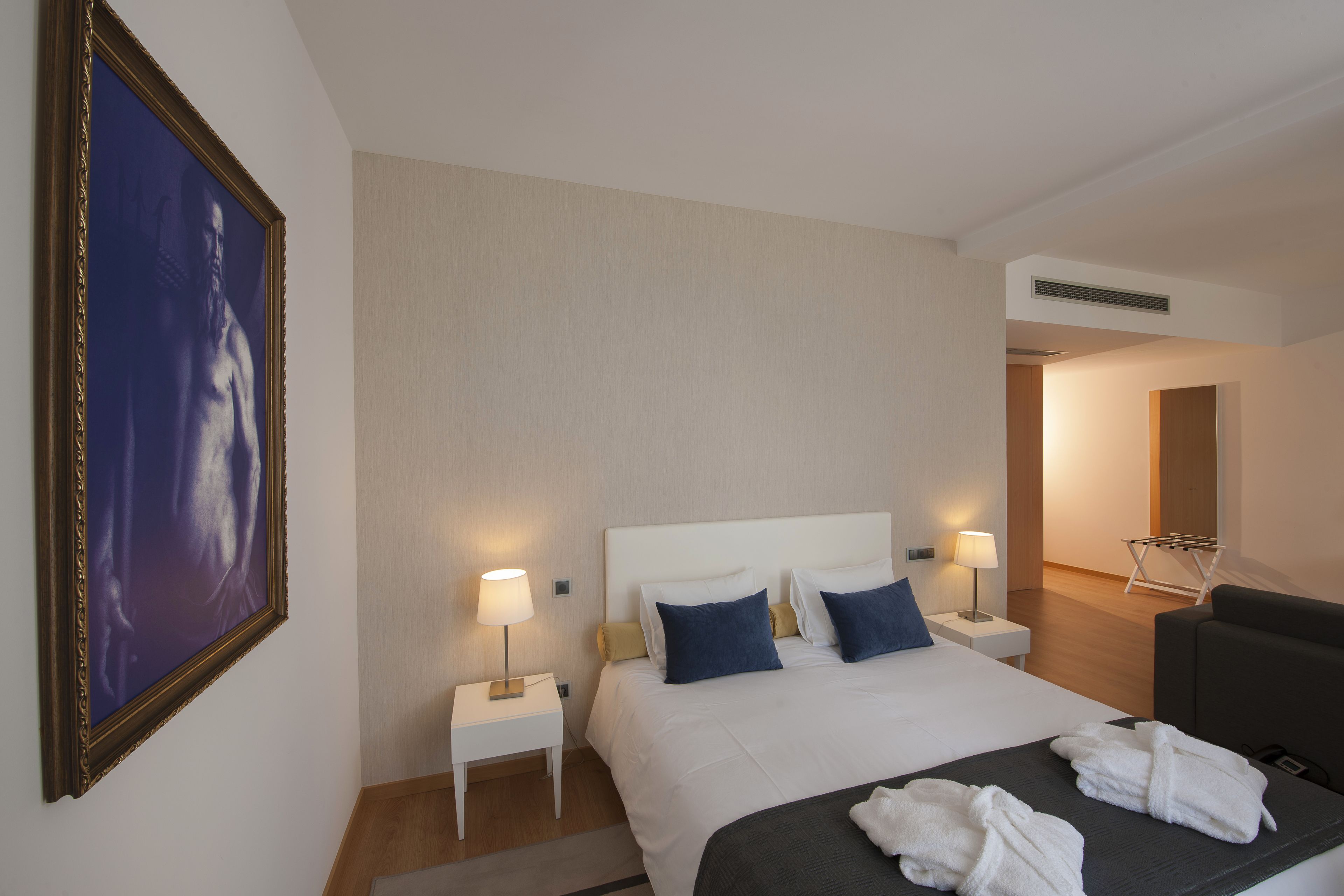 A hotel room with a large bed, two bedside tables with lamps, a dark-colored sofa, and a framed picture on the wall.