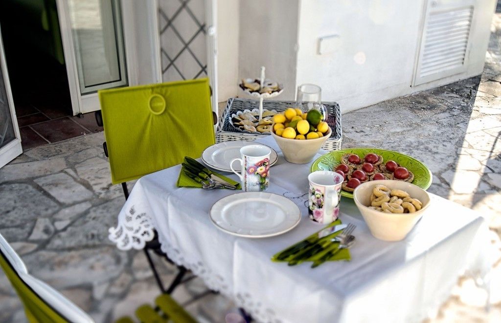 Mesa posta com pratos, copos e tigelas de comida, incluindo limões e azeitonas, em um terraço com piso de pedra.