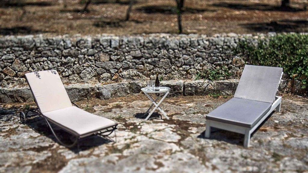 Due lettini con struttura in metallo e cuscini, un piccolo tavolino rotondo con base metallica e un muro di pietra sullo sfondo.