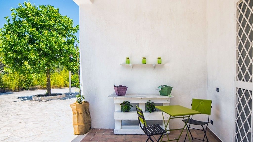 A patio with a white wall, a green table, and two chairs. There are potted plants on the wall and a large pot on the ground.