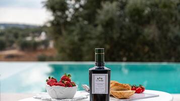 A bottle of L'Iconic olive oil, a bowl of strawberries, and a plate of bread by a poolside table.