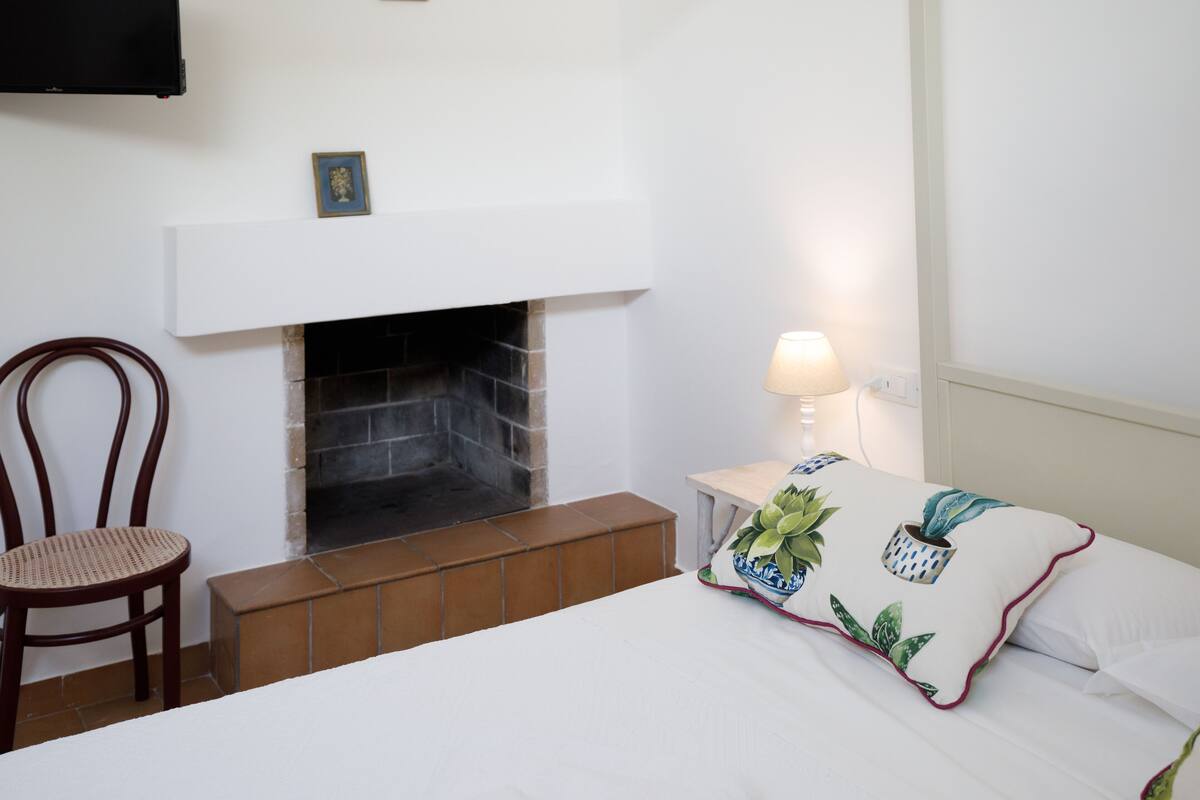 A bedroom with a fireplace, a bedside table with a lamp, a chair, and a bed with a floral pillow.