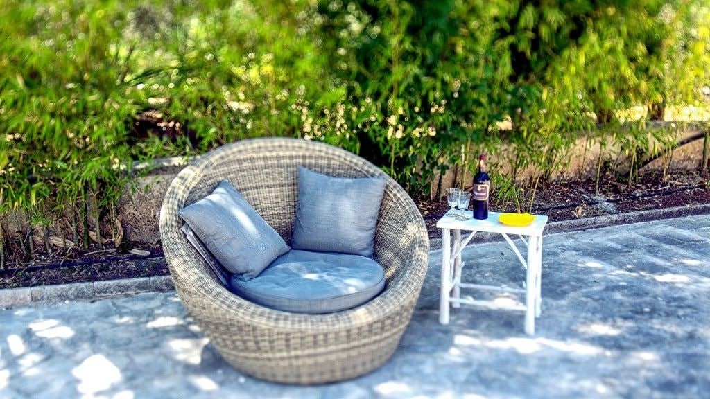 A wicker chair with cushions, a small white table with a bottle and glasses, and a yellow plate.