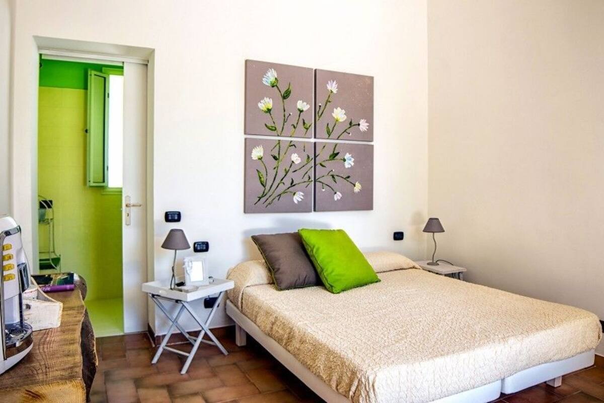 A bedroom with a bed, a desk with a lamp, a coffee machine, and a painting on the wall.