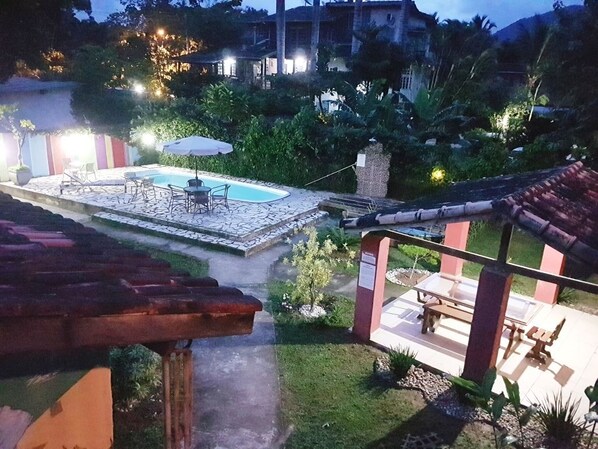 Zona de piscina con sillas de descanso y una mesa, rodeada de vegetación y un camino de piedra.