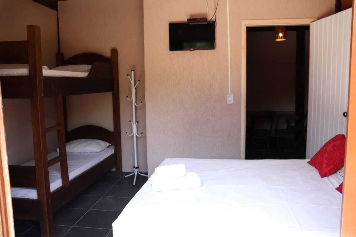 A room with a bunk bed, a single bed with a red pillow, a coat rack, and a television mounted on the wall.