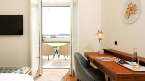 A hotel room with a bed, a desk with a lamp, a chair, and a view of a garden and lake through an open door. - Montebelo Vista Alegre Ílhavo Hotel (Ilhavo)