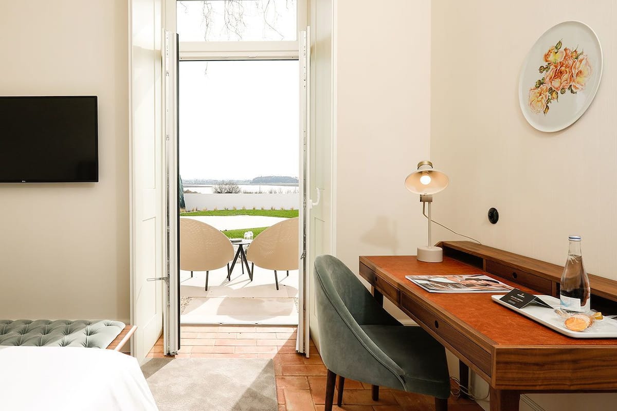 A hotel room with a bed, a desk with a lamp, a chair, and a view of a garden and lake through an open door.