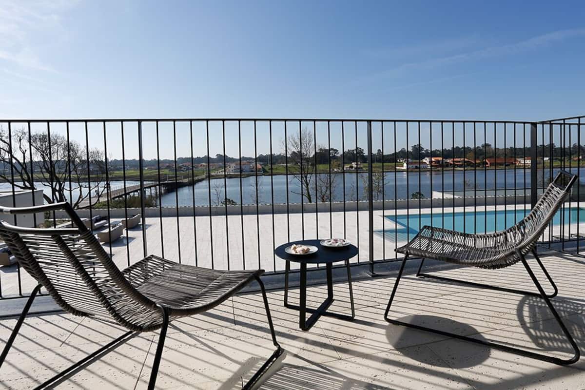 A deck with two lounge chairs and a small table, overlooking a swimming pool and a body of water.