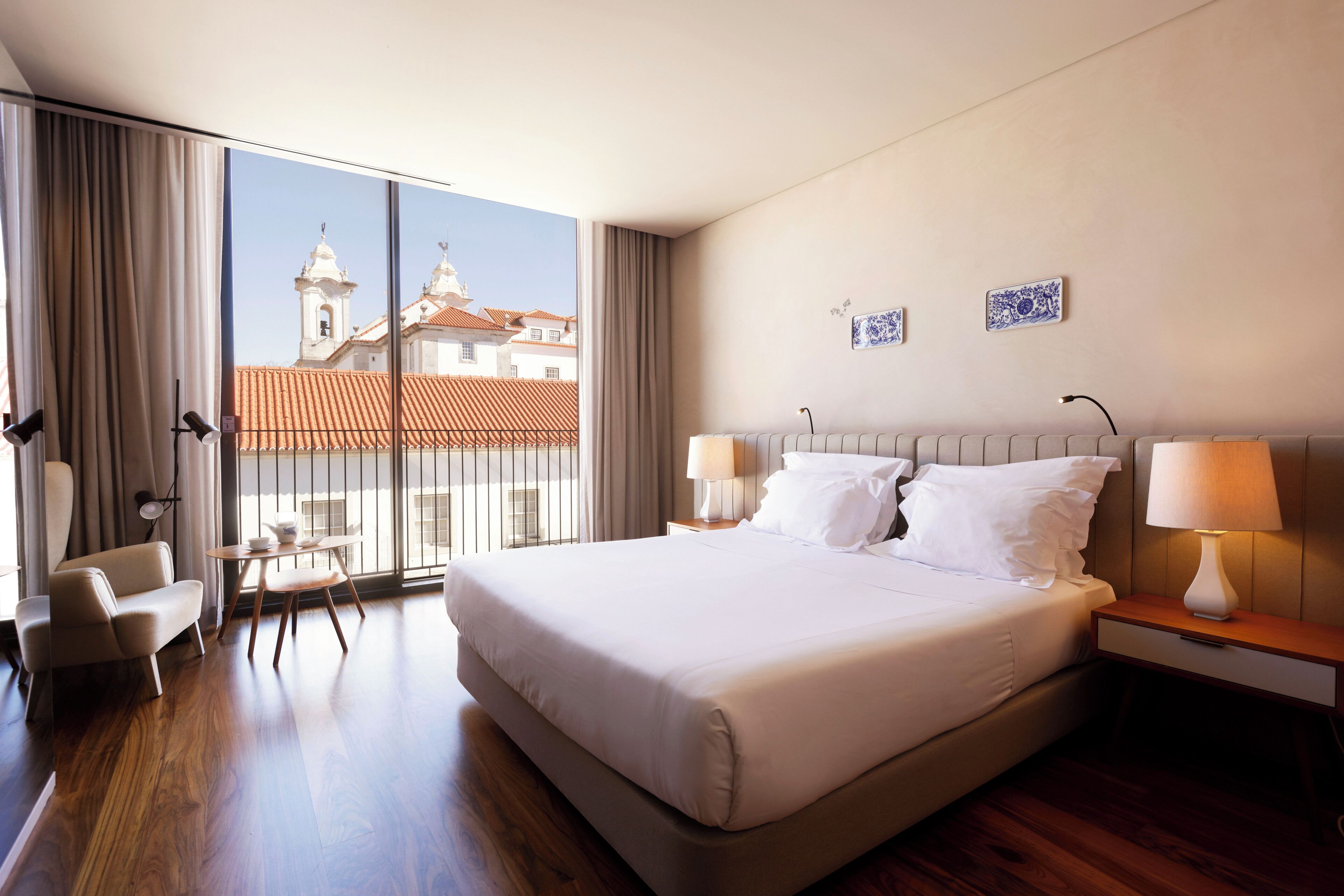 Quarto de hotel com uma cama grande, duas mesas de cabeceira com luminárias, uma cadeira, uma pequena mesa e vista para edifícios através de janelas amplas.