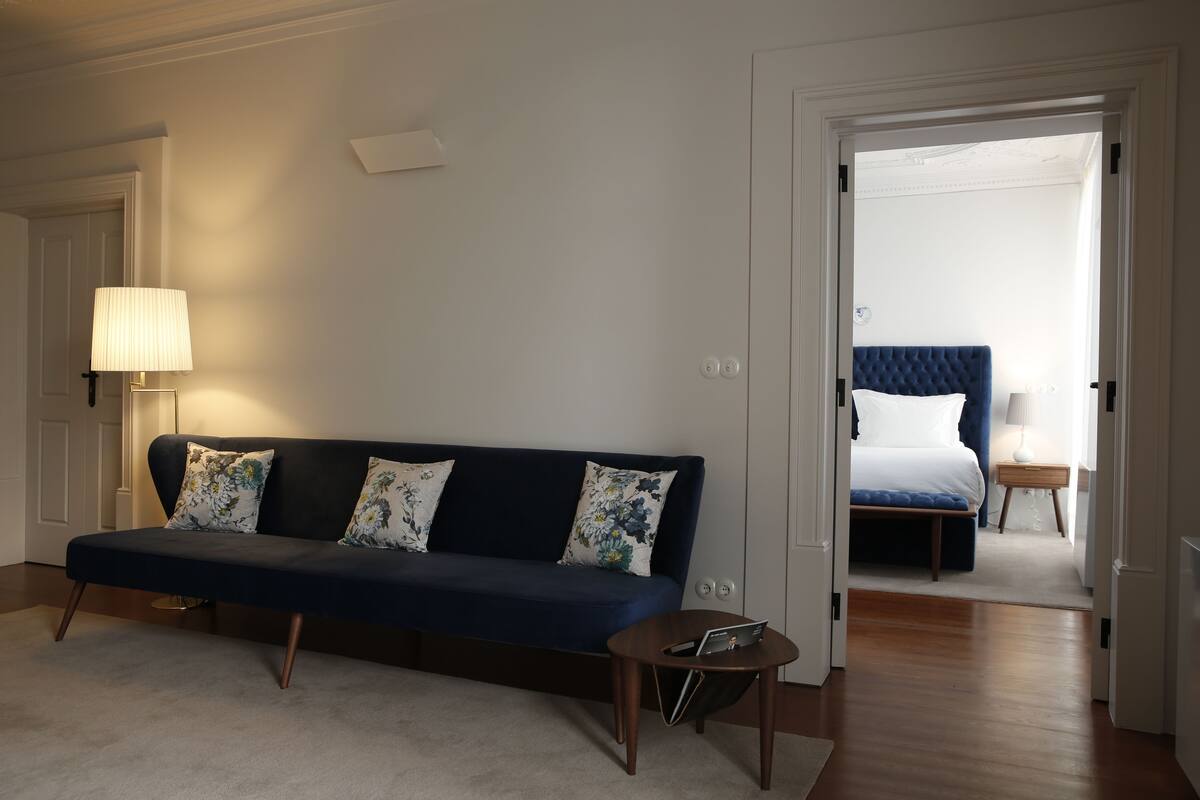 A room with a dark blue sofa, a small round table, and a lamp. There is a door leading to a bedroom with a bed and a nightstand.