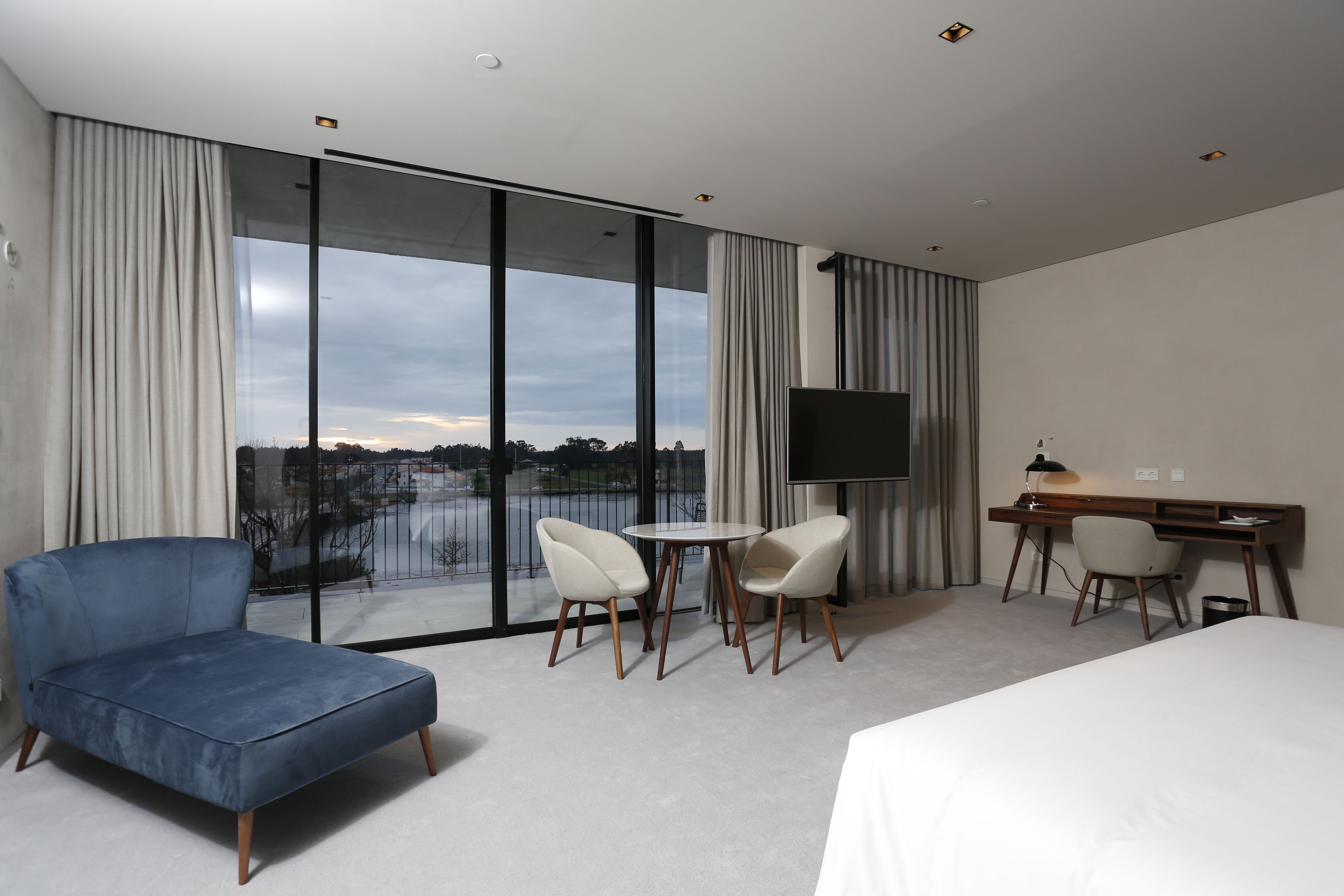 A modern hotel room with a large bed, a desk with a chair, a small round table with two chairs, and a TV.