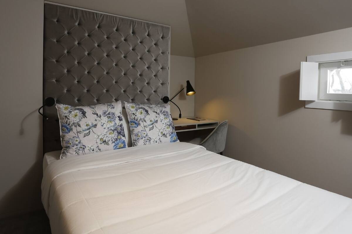 A bedroom with a tufted headboard, a bed with white bedding, a desk with a lamp, and a TV mounted on the wall.