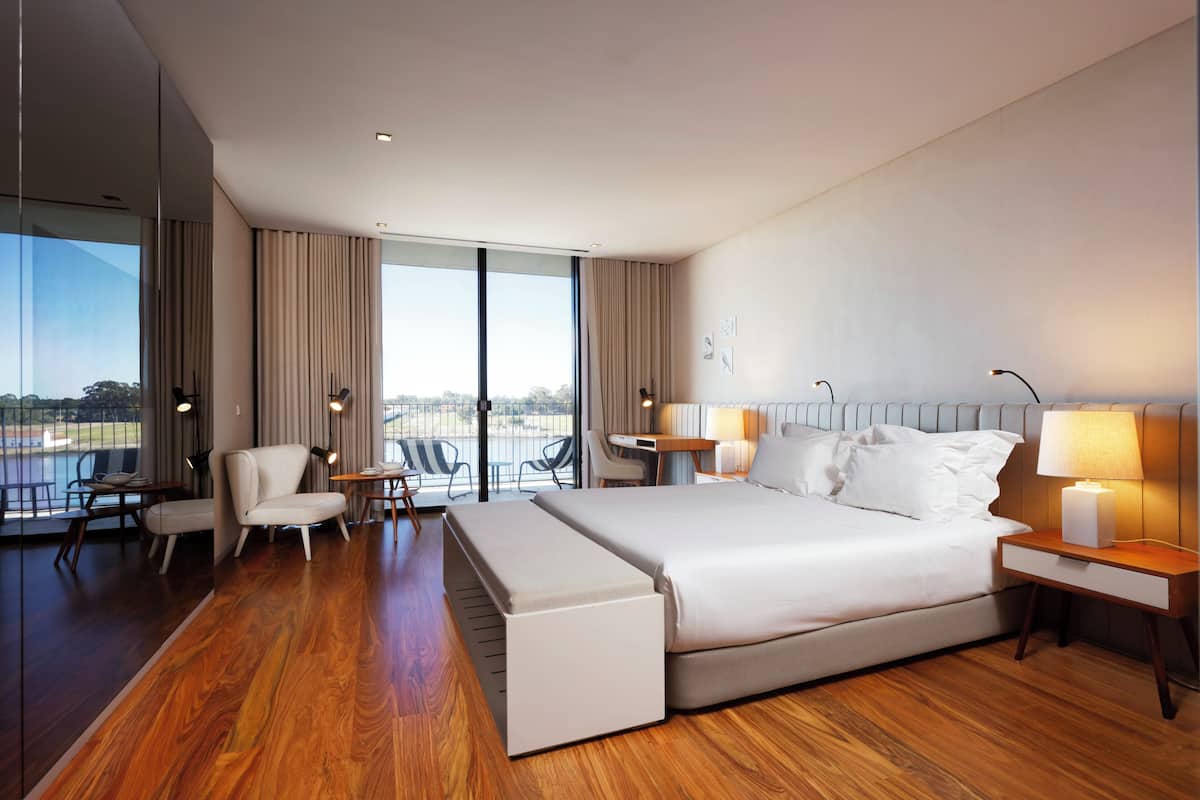 A modern hotel room with a large bed, a desk, a chair, and a view of the outdoors.