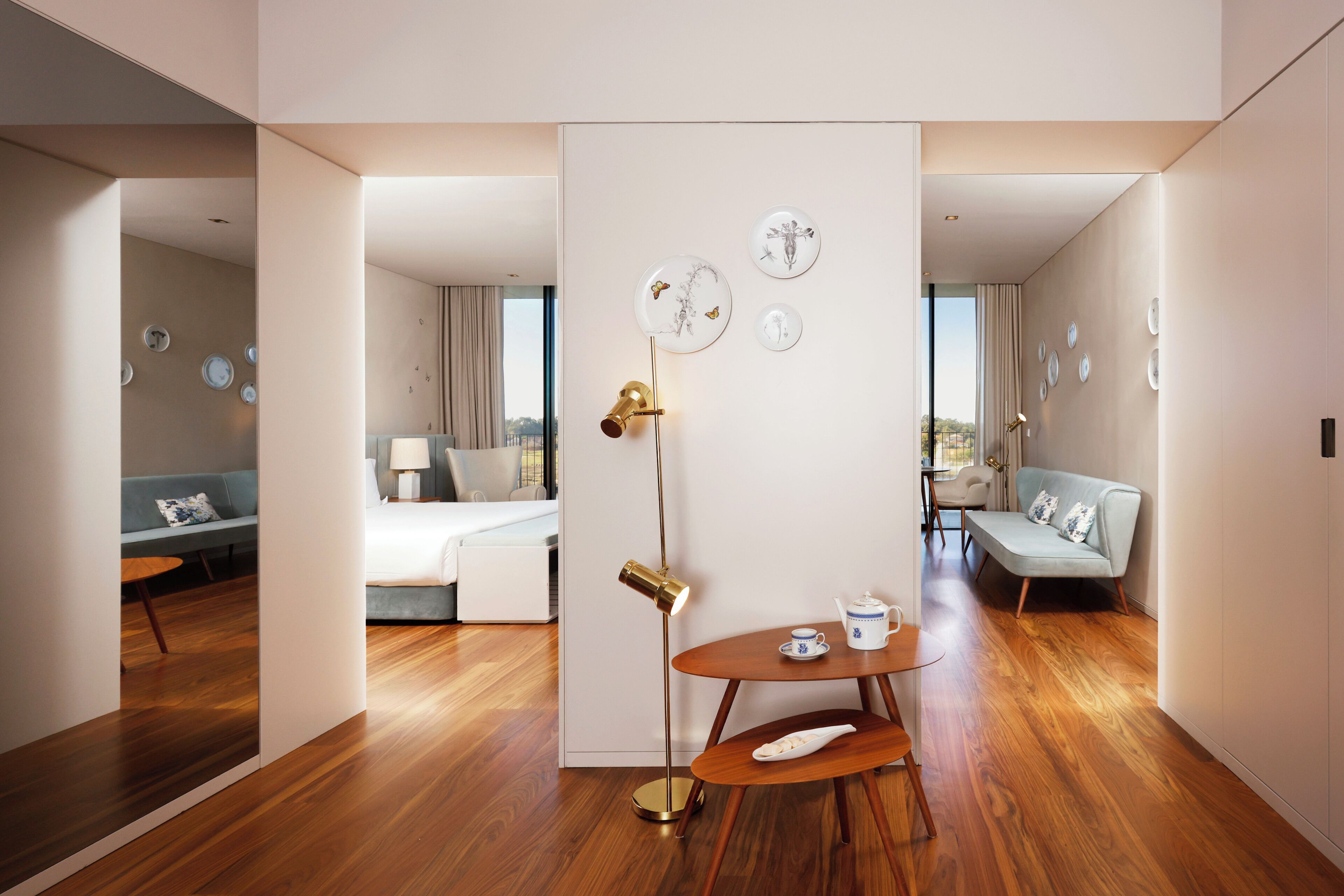 A modern hotel room with a wooden floor, a round table with a teapot, a sofa, and a bed.
