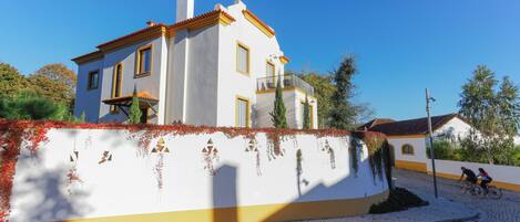 Un edificio de dos pisos con fachada blanca y detalles amarillos, una calle adoquinada y un cielo azul despejado.