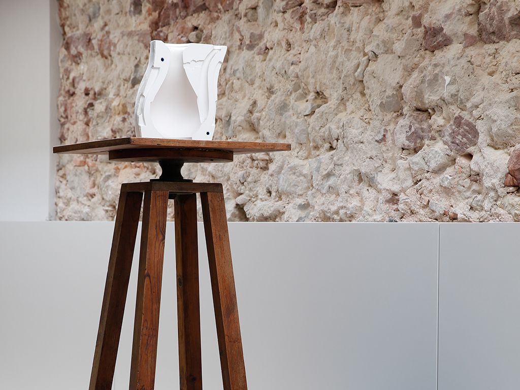 Una escultura blanca sobre un pedestal de madera frente a una pared con textura.