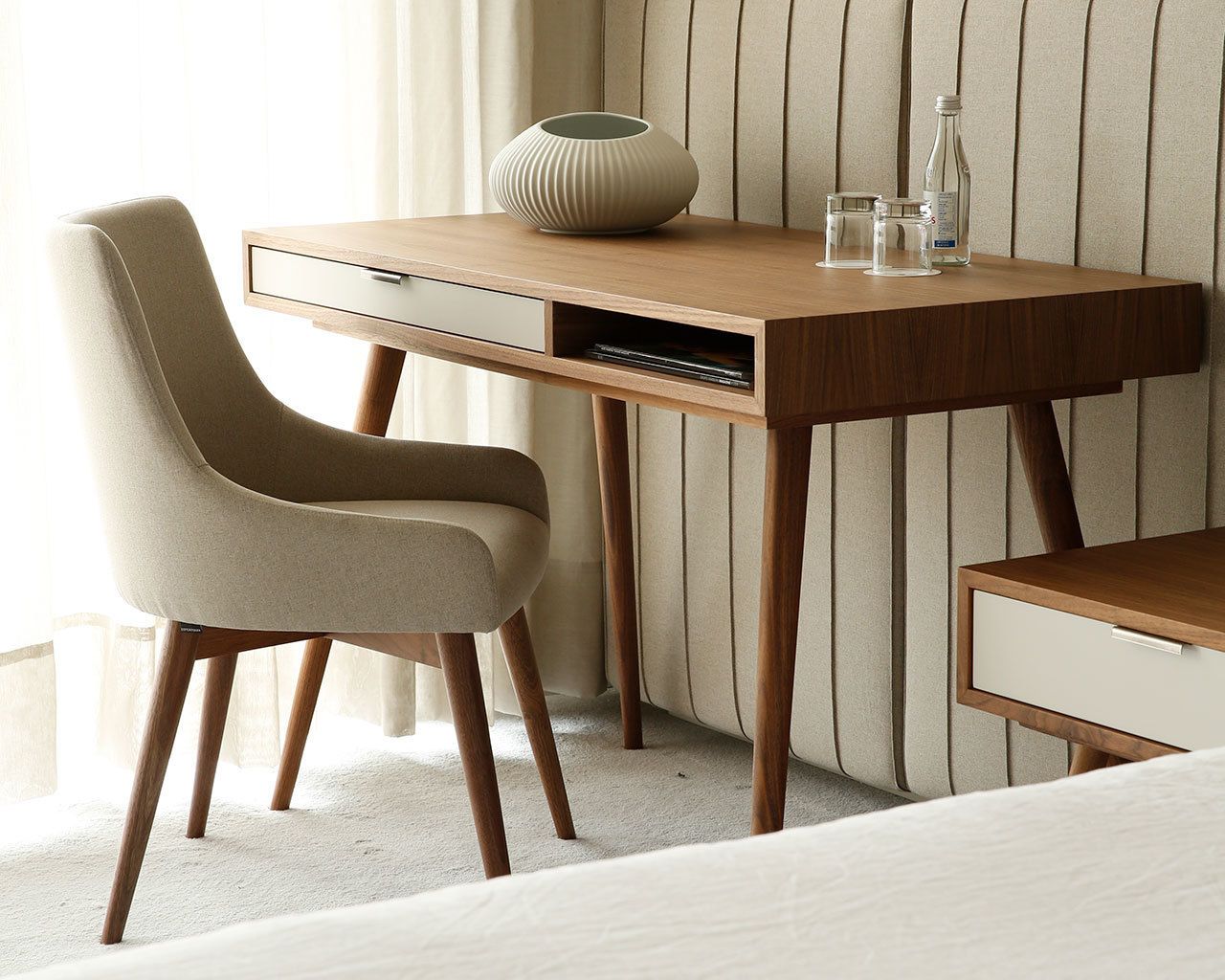A hotel room with a wooden desk, a beige chair, a vase, and a bottle on the desk.