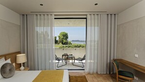 A hotel room with a bed, a desk, a chair, a small table with chairs, and a view of a garden and water body through a large window. - Montebelo Vista Alegre Ílhavo Hotel (Ilhavo)