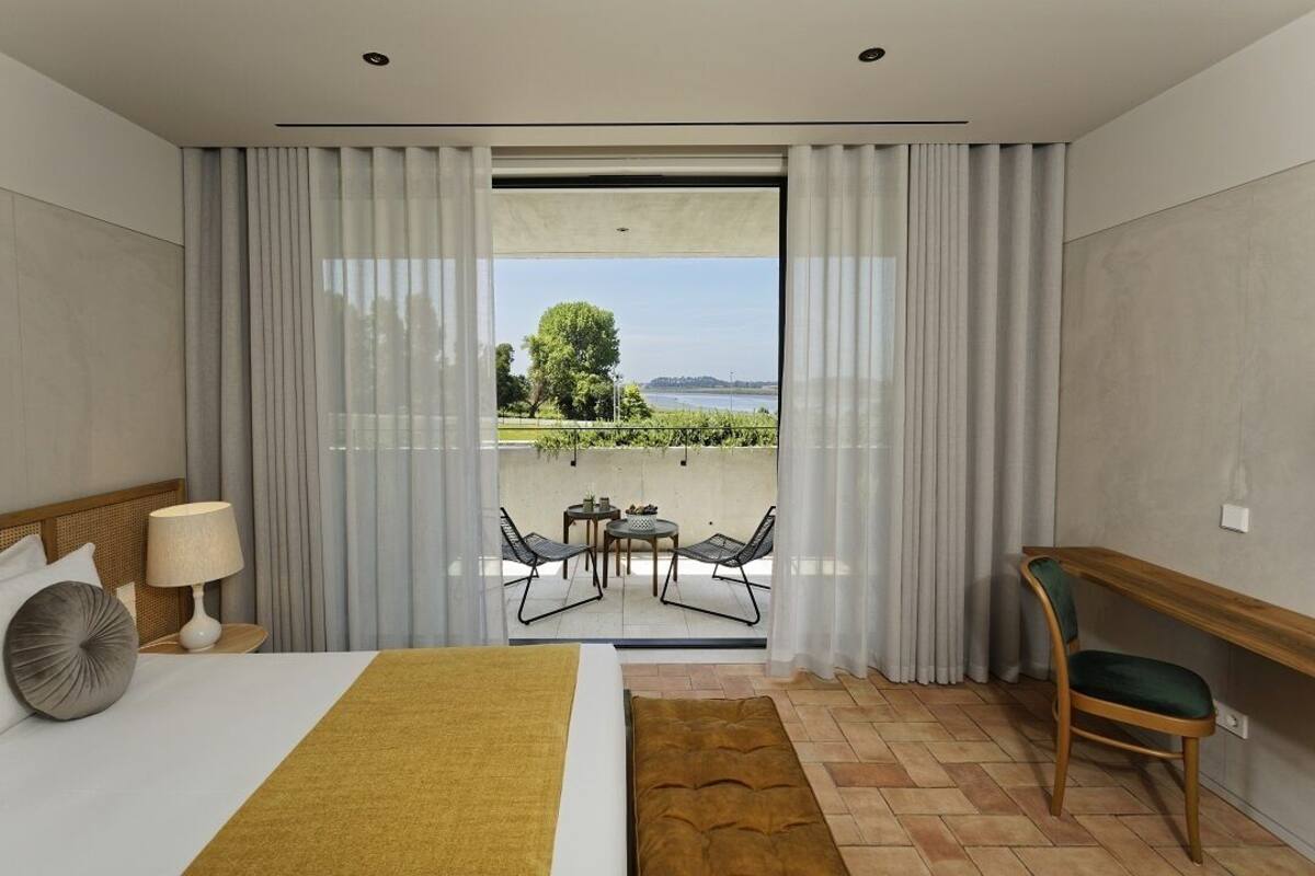 A hotel room with a bed, a desk, a chair, a small table with chairs, and a view of a garden and water body through a large window.