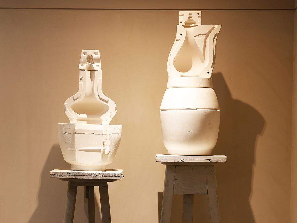 Two white ceramic sculptures on wooden pedestals against a plain wall.