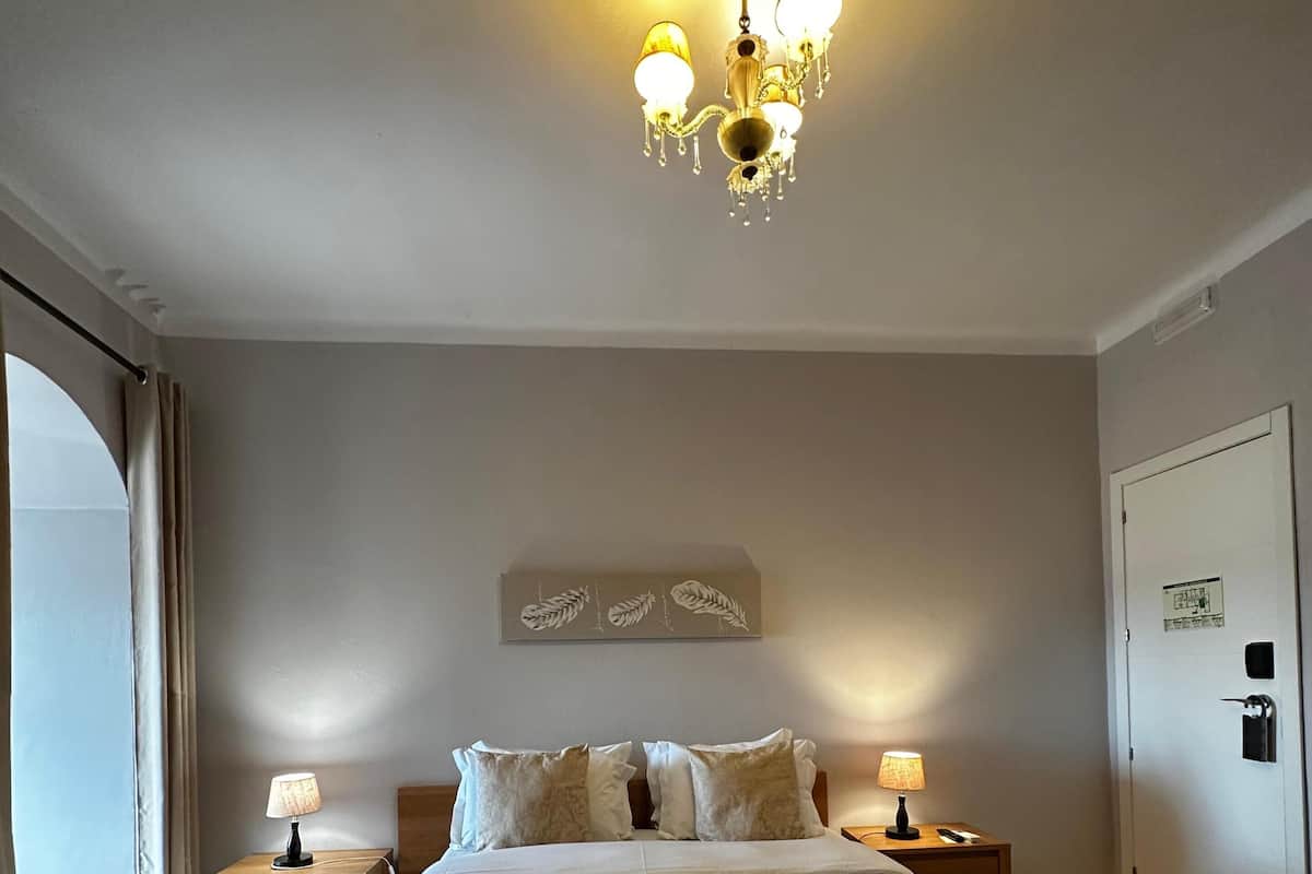 A hotel room with a large bed, two bedside tables with lamps, a chandelier, a door with a lock, and a window with curtains.