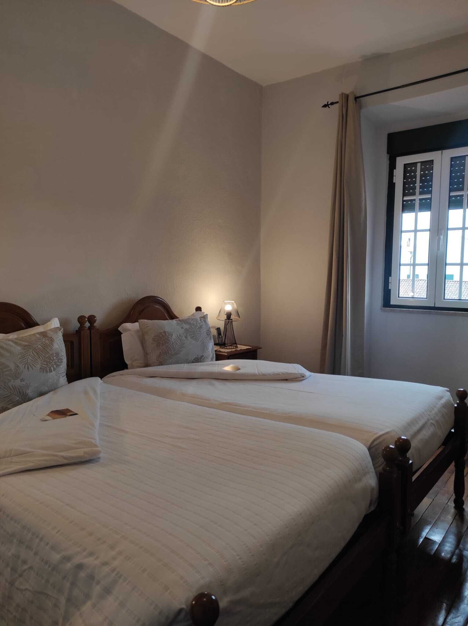 Una habitación con dos camas, una mesita de noche con lámpara, una ventana con cortinas y suelo de madera.