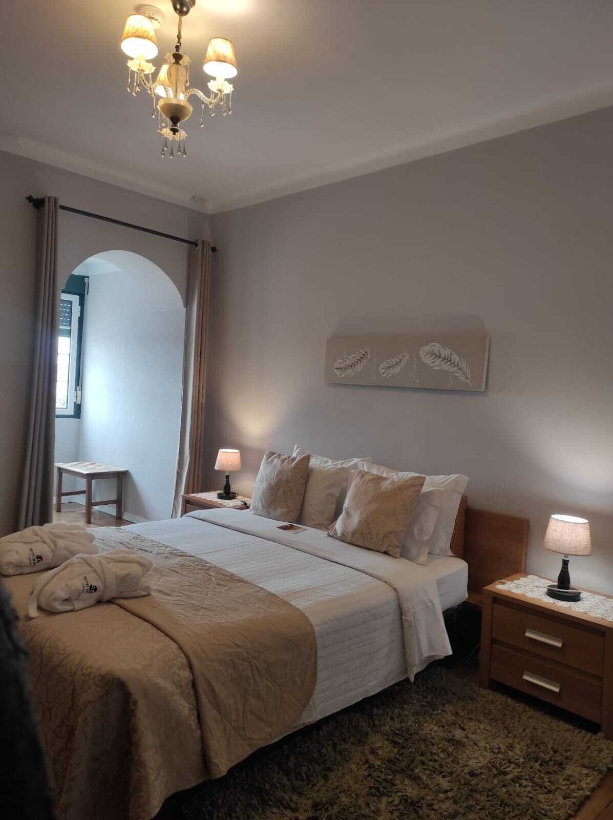 A bedroom with a large bed, two bedside tables with lamps, a chandelier, a mirror, and a wall-mounted artwork.