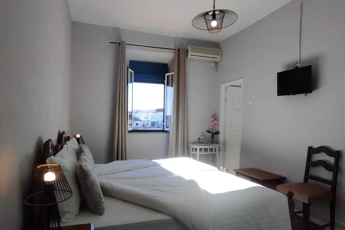 A bedroom with a bed, a bedside lamp, a chair, a TV, and a window with curtains.