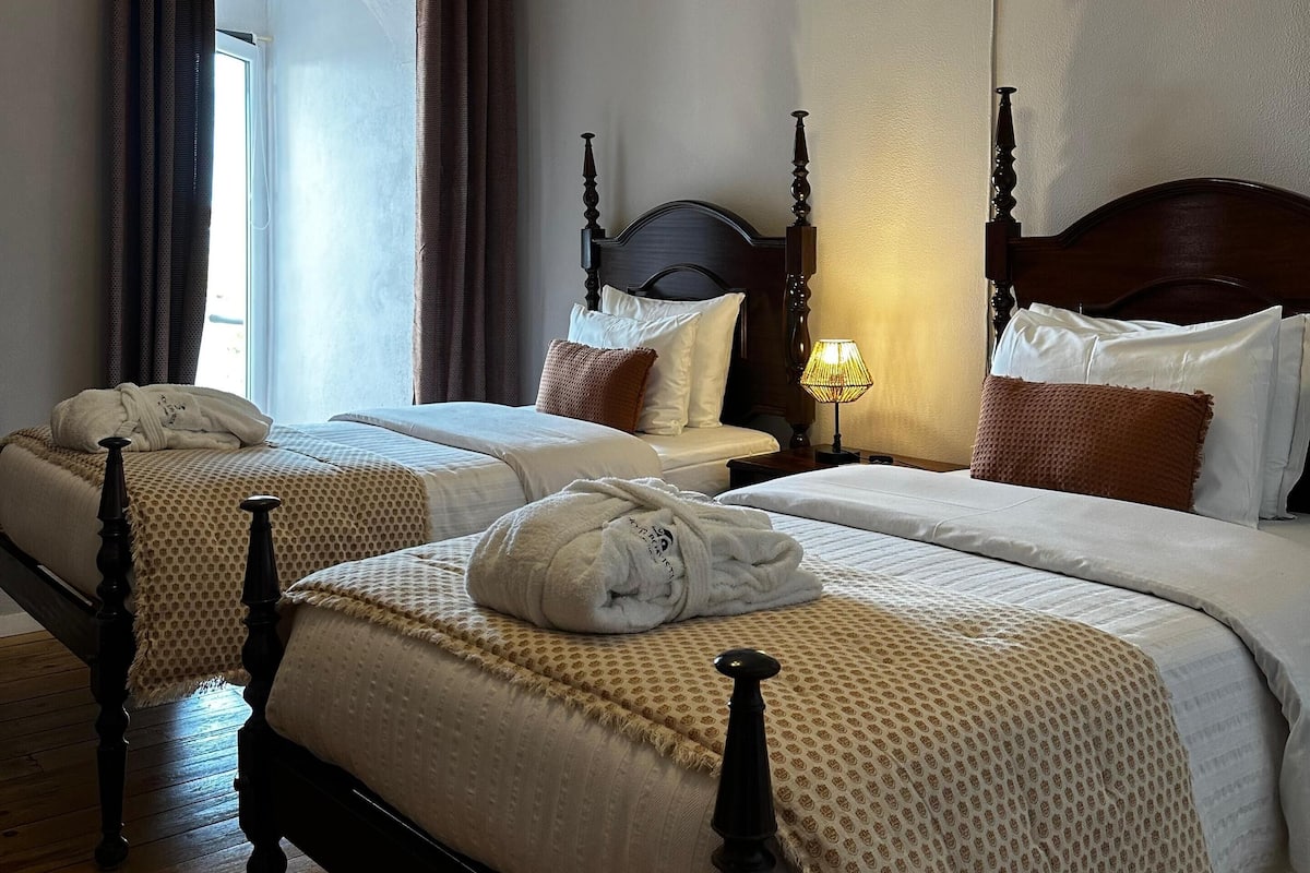 A hotel room with two beds, a chandelier, and a view of the outdoors through a window with curtains.