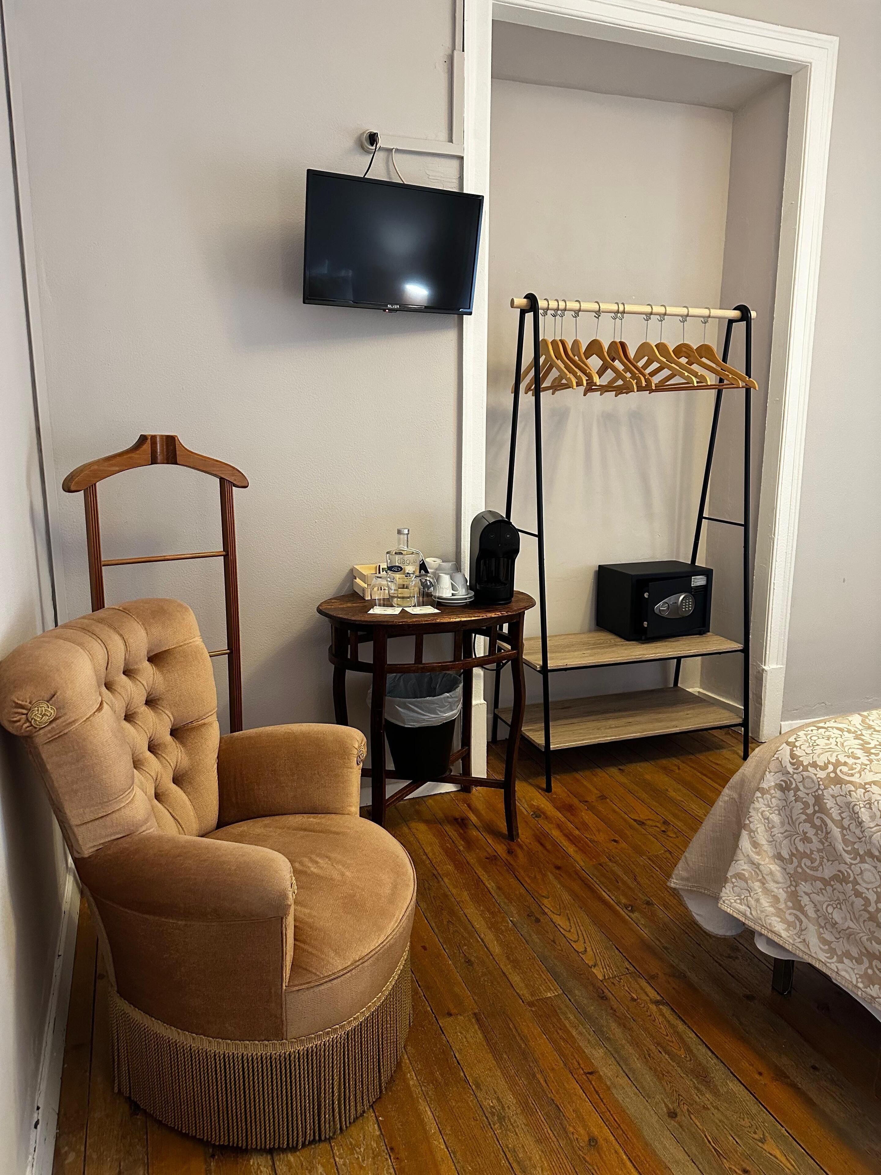 A room with a tufted armchair, a small table with toiletries, a hanging clothes rack, a television, and a bed with a patterned bedspread.